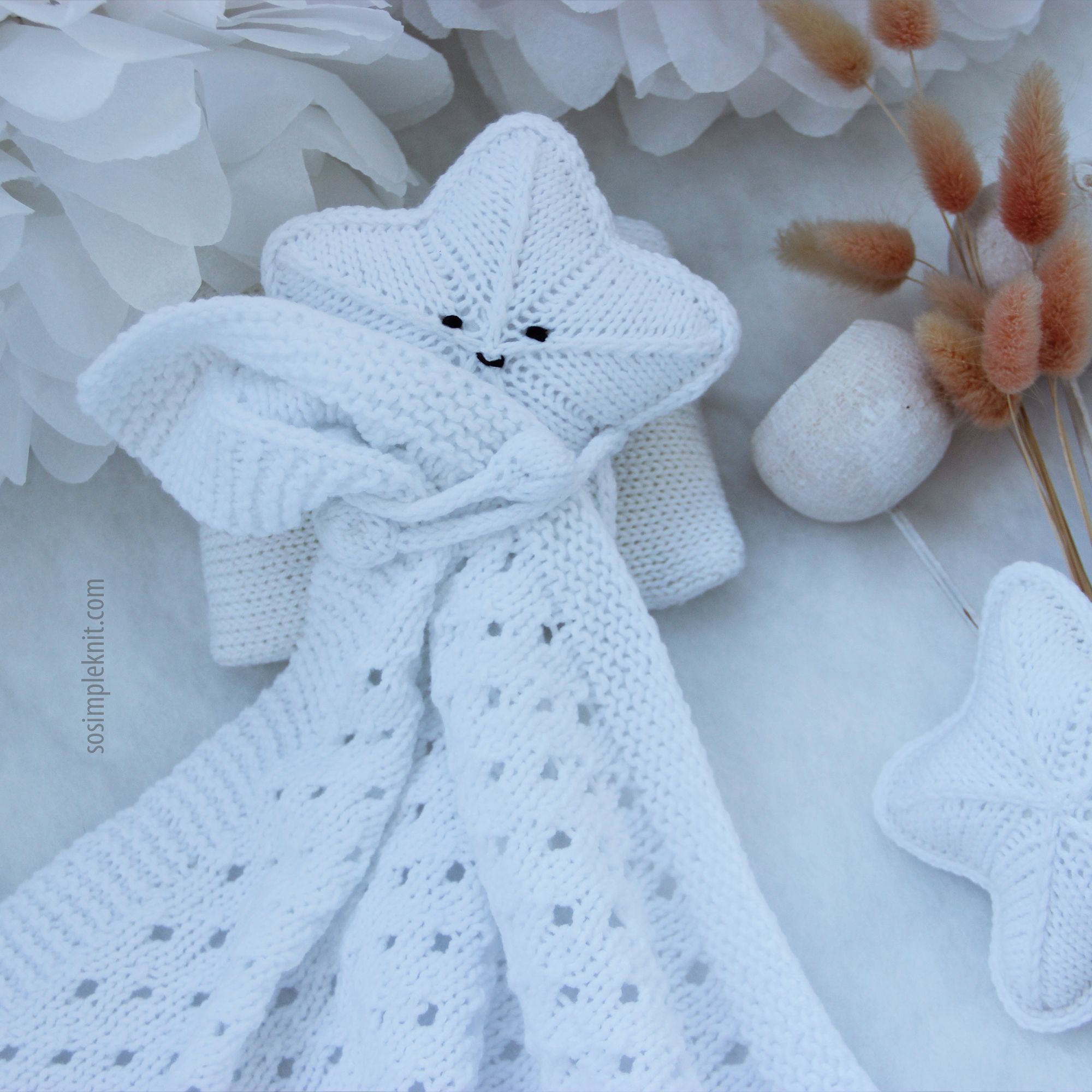 A white knitted security blanket shaped like a star, featuring a smiling face and stitched eyes, is positioned against a light background. The blanket is intricately patterned with openwork and textured stitches, creating a soft, cozy appearance. Surrounding the blanket are neutral-toned decorative elements, including dried flowers and a small pom-pom, adding a gentle touch to the scene. The overall composition is serene and inviting, emphasizing the softness and charm of the blanket.