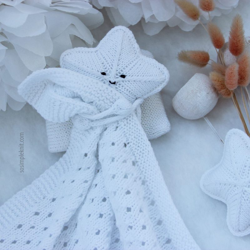 A white knitted security blanket shaped like a star, featuring a smiling face and stitched eyes, is positioned against a light background. The blanket is intricately patterned with openwork and textured stitches, creating a soft, cozy appearance. Surrounding the blanket are neutral-toned decorative elements, including dried flowers and a small pom-pom, adding a gentle touch to the scene. The overall composition is serene and inviting, emphasizing the softness and charm of the blanket.