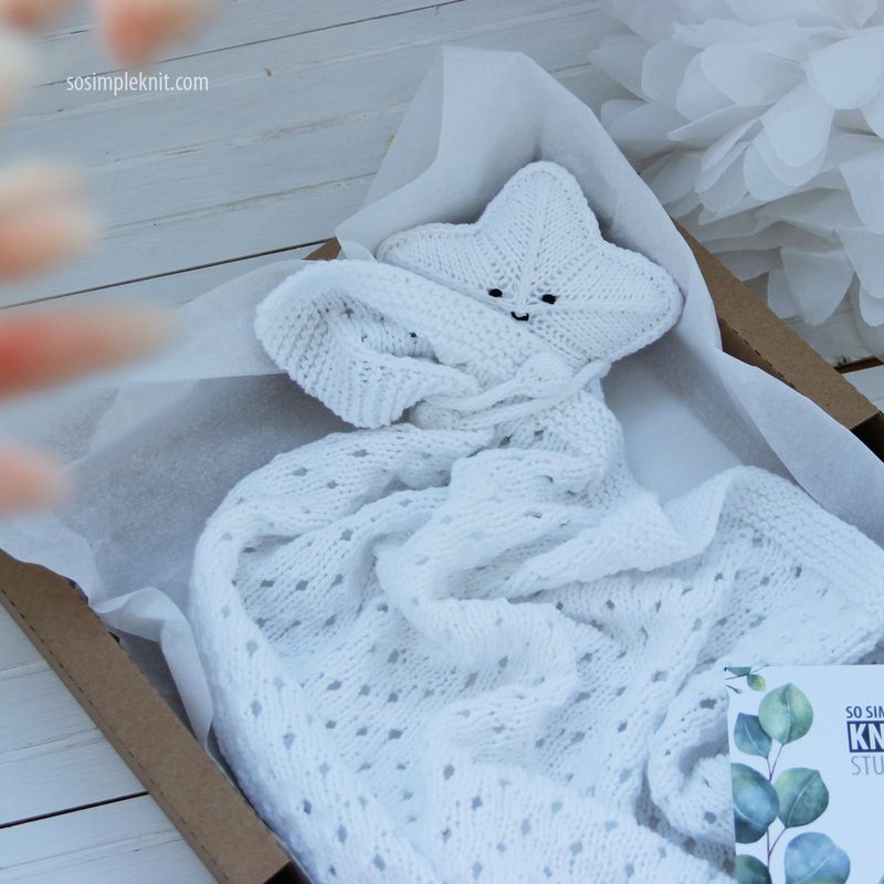 A knitted security blanket in the shape of a star, featuring a soft, white texture and a smiling face. The blanket is neatly folded inside a brown box, with a delicate lace pattern visible on the fabric. Surrounding the box are light-colored decorative elements, including tissue paper and a flower, adding a soft, whimsical touch to the scene. The image has a bright and airy feel, emphasizing the warmth and charm of the handmade item.