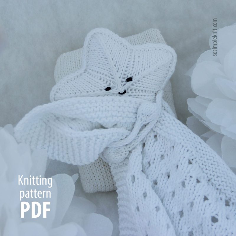 The image features a white knitted security blanket shaped like a star, with a smiling face made from black buttons stitched onto the star's head. The blanket's body has a textured, lacy pattern, and it is tied at the base of the star, creating a soft drape. The background consists of soft white fabric and large white paper flowers, creating a serene and peaceful presentation. The text on the left side reads "Knitting pattern PDF" in a simple font.