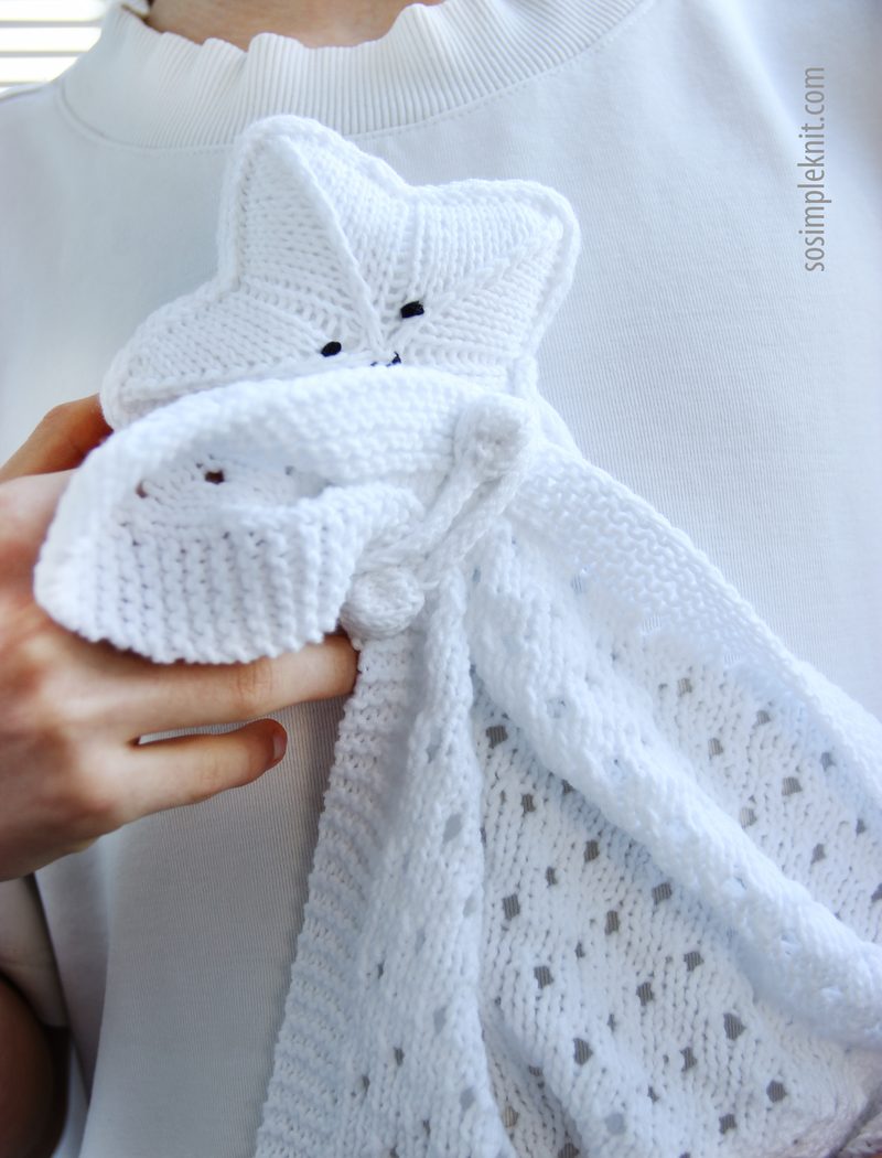 A person holds a white knitted security blanket shaped like a star, with a textured, lace-like design on the blanket portion. The star has small, black button eyes and a smiling mouth, contrasting with the white fabric. The person's hand and part of a white shirt are visible in the frame, focusing on the blanket. The background is neutral and uncluttered, emphasizing the blanket's details.