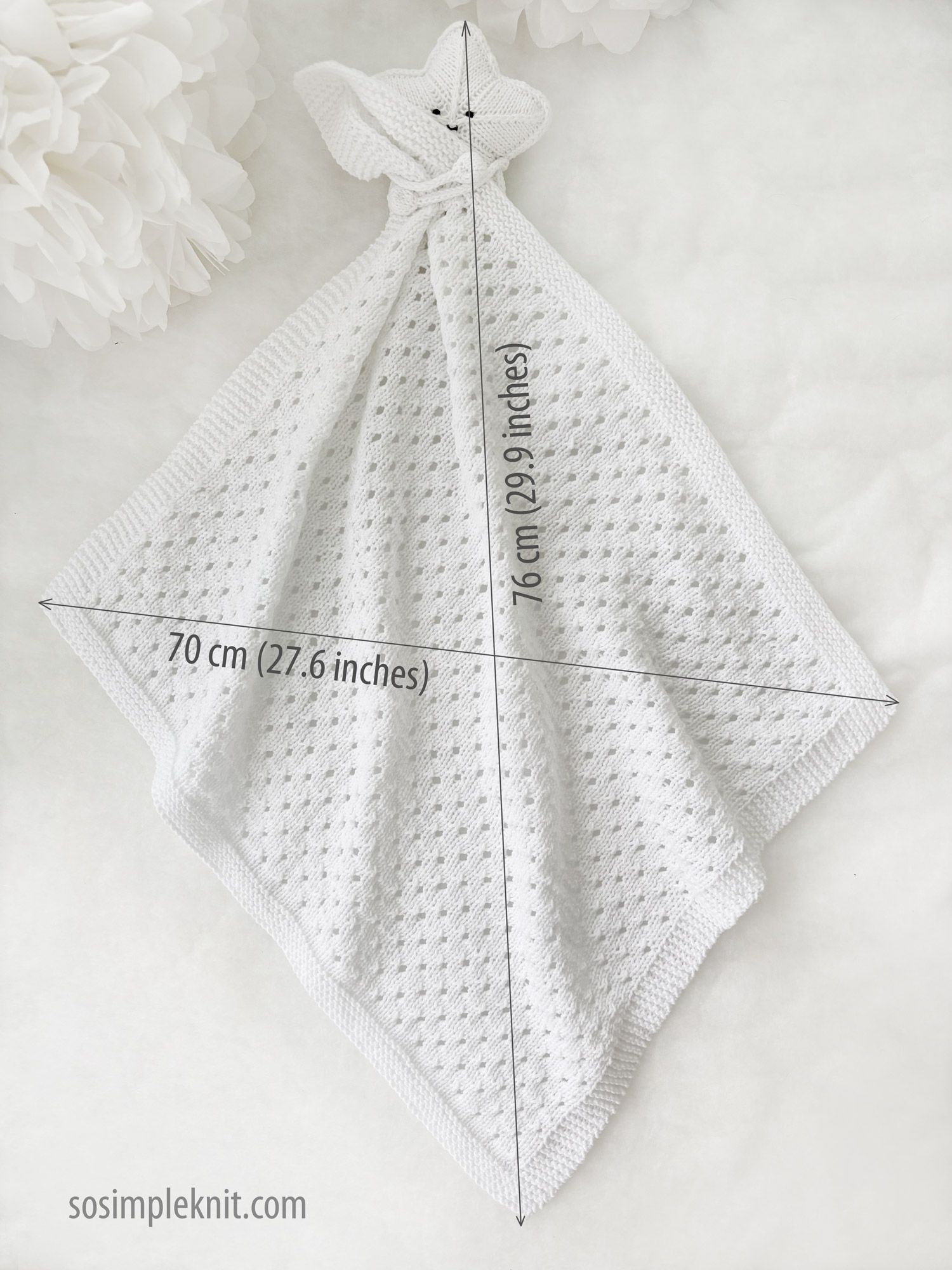 A white knitted security blanket shaped like a star, featuring a textured stitch pattern and a hood resembling a teddy bear's face with two small fabric ears. The blanket is displayed diagonally, with measurements annotated in the corners: 70 cm (27.6 inches) by 76 cm (29.9 inches). The background is light and neutral, enhancing the softness and simplicity of the blanket's design.