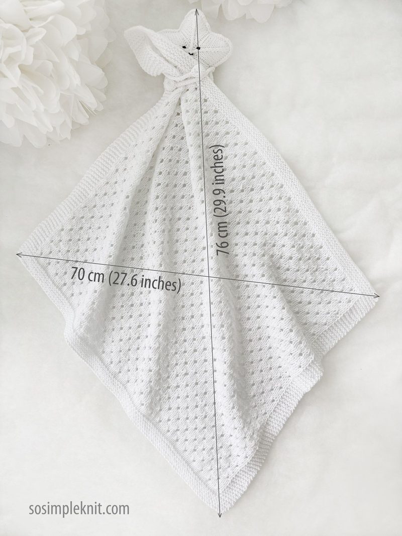 A white knitted security blanket shaped like a star, featuring a textured stitch pattern and a hood resembling a teddy bear's face with two small fabric ears. The blanket is displayed diagonally, with measurements annotated in the corners: 70 cm (27.6 inches) by 76 cm (29.9 inches). The background is light and neutral, enhancing the softness and simplicity of the blanket's design.