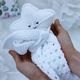 A pair of hands gently holds a white knitted security blanket shaped like a star, featuring a smiling face with two black button-like eyes. The star's body has a textured knit pattern, while the blanket has a lacy design along the edges. The background is softly blurred, with light colors and floral motifs, creating a serene, cozy atmosphere.