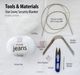 The image shows a flat lay of materials for the "Star Lovey Security Blanket" knitting pattern on a light background. It includes a ball of white cotton yarn labeled "YarnArt jeans," a long circular knitting needle, stuffing, flexible needles, scissors, a measuring tape, stitch markers, a tapestry needle, and some paper clips. The items are arranged neatly, with labels placed at the top indicating "Tools & Materials." The overall color palette is predominantly white with hints of pastel.