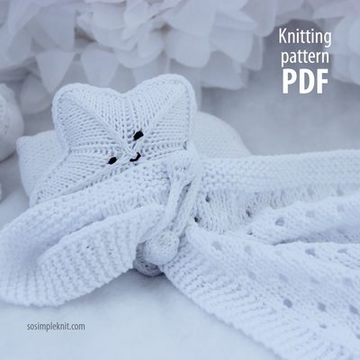 A white knitting pattern for a star-shaped security blanket is displayed, showcasing a textured star head with stitched facial features. The blanket has a long, flat body that elegantly drapes, with a knot at the bottom. The background is light and blurred, featuring soft white petals, giving a gentle and serene feel to the image. The text "Knitting Pattern PDF" is positioned in the upper right corner in a clean, modern font.