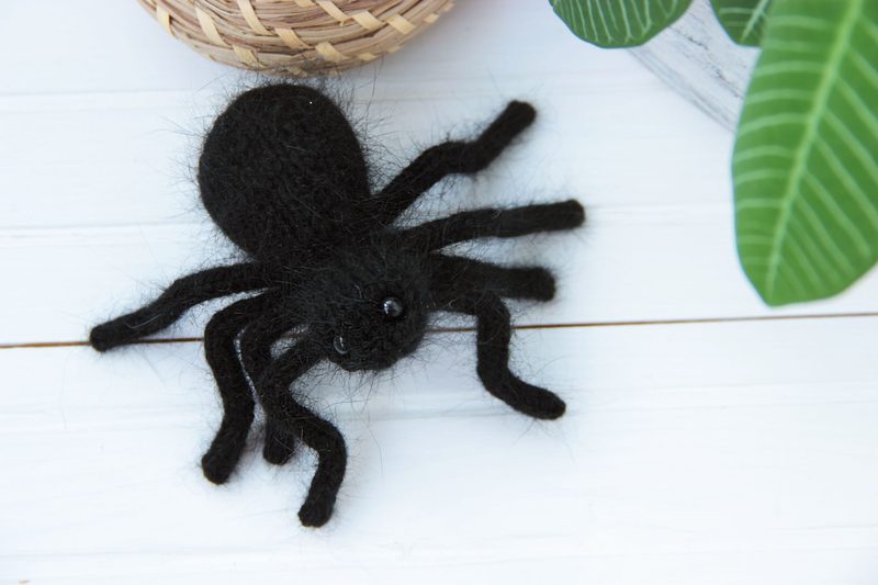 A black knitted tarantula toy is positioned on a white wooden surface, showcasing its eight legs and round body. The spider has glossy eyes, enhancing its playful appearance. In the background, a woven basket and a green plant with large leaves are partially visible, providing a contrasting texture and color.