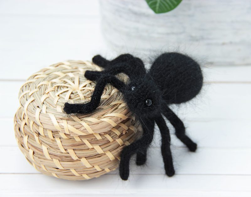 A black knitted toy tarantula sits atop a woven basket made of natural fibers. The spider has a round body and long, fuzzy legs, showcasing a textured appearance. It is positioned slightly off-center, with the basket providing a contrasting light brown color against the dark tarantula. The background features a blurred green plant in a textured pot, adding a hint of color without distracting from the main subject. The overall composition emphasizes the craftsmanship of the knitted spider.