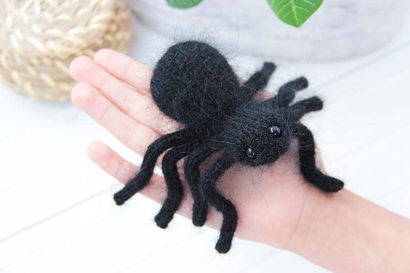 A handmade black tarantula toy is resting on an outstretched hand, showcasing its fuzzy texture and prominent round body. The spider has eight long, curved legs and two shiny black eyes. The background features a softly blurred natural setting with green foliage and a woven basket, enhancing the focus on the spider. The image is brightly lit, emphasizing the contrast between the black spider and the light-colored hand.