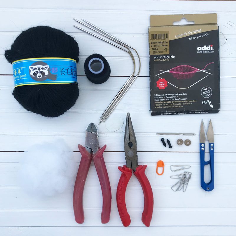 The image features a black skein of yarn with a raccoon label, a spool of black thread, knitting needles, a pack of addi knitting tools, a pair of red pliers, a small orange tool, a few black beads, safety pins, and a pair of scissors, all arranged on a white wooden surface.