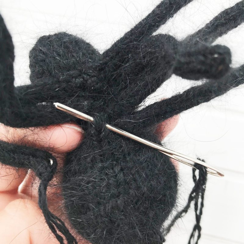 A close-up image of a black knitted tarantula, with textured yarn forming its body and long, thin legs. A stainless steel sewing needle is visible, partially inserted into the body, indicating the finishing process. The background is light and neutral, providing contrast to the black spider. The overall focus is on the intricate details of the knitting.