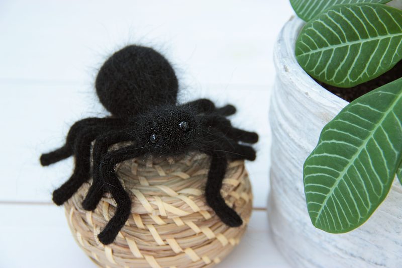 A black knitted tarantula with fuzzy details sits atop a light wicker basket, featuring eight prominent legs and glossy eyes. To the right, there is a green potted plant with glossy, textured leaves and a light grey, textured pot. The background is a soft, white wooden surface, providing a neutral contrast to the dark spider and vibrant greenery.