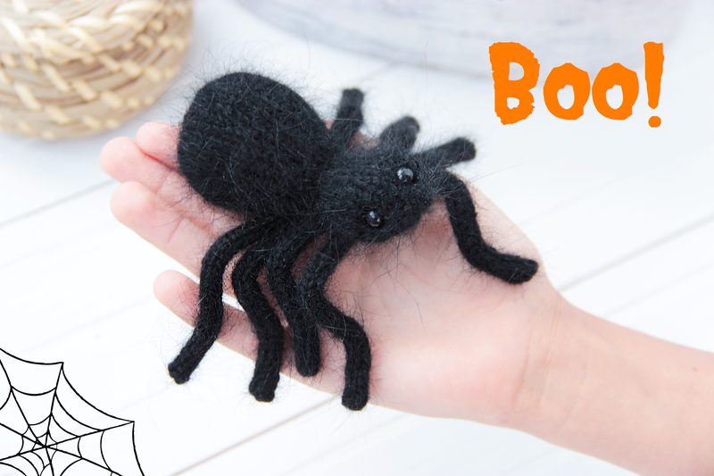 A hand holds a small, black, felted spider with eight fuzzy legs and glossy plastic eyes, set against a light background. The spider's body is round and fuzzy, while the legs are long and slightly curved. In the top right corner, the word "Boo!" is displayed in an orange, bold font. A spider web illustration is in the bottom left corner of the image, adding a Halloween theme. The overall style is playful and suitable for a toy design.