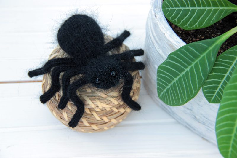 A small, handmade black tarantula toy sits atop a woven basket, featuring eight legs and a rounded body with a fluffy texture. The tarantula has two shiny black eyes and is positioned on a light wooden surface, with a potted plant visible in the background, showcasing large green leaves in a textured gray pot. The overall color palette is dark and natural, emphasizing the spider's details against the lighter background.