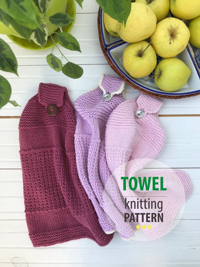Three hanging kitchen towels in varying shades of pink and purple are displayed on a white wooden surface. The towels have a textured pattern with buttons for hanging. To the left, there is a green plant with broad leaves and to the right, a blue bowl filled with yellow apples. The text "TOWEL knitting PATTERN" is overlaid on the image in bold green and gray letters.