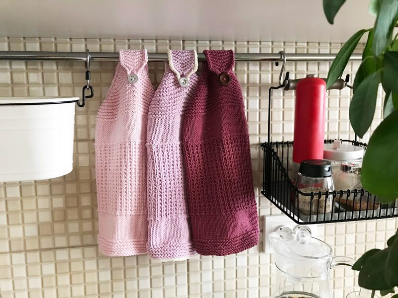 Three knitted hanging kitchen towels in varying shades of pink—light pink, medium pink, and a deep magenta—are displayed on a metal rod. Each towel features a button closure at the top. The background consists of a light-colored tiled wall, and there is a black wire basket containing kitchen items visible to the right, alongside a red bottle and a white pot.