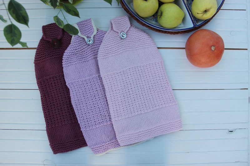 The image features three hanging kitchen towels displayed on a light wooden surface, with the towels arranged in a row. The left towel is a deep burgundy with a button detail, while the middle and right towels are both shades of light pink, showcasing textured knit patterns. A bowl of green apples and a single orange are positioned to the side of the towels, providing a contrasting pop of color against the soft hues of the fabrics. The overall composition is well-lit and aesthetically pleasing.