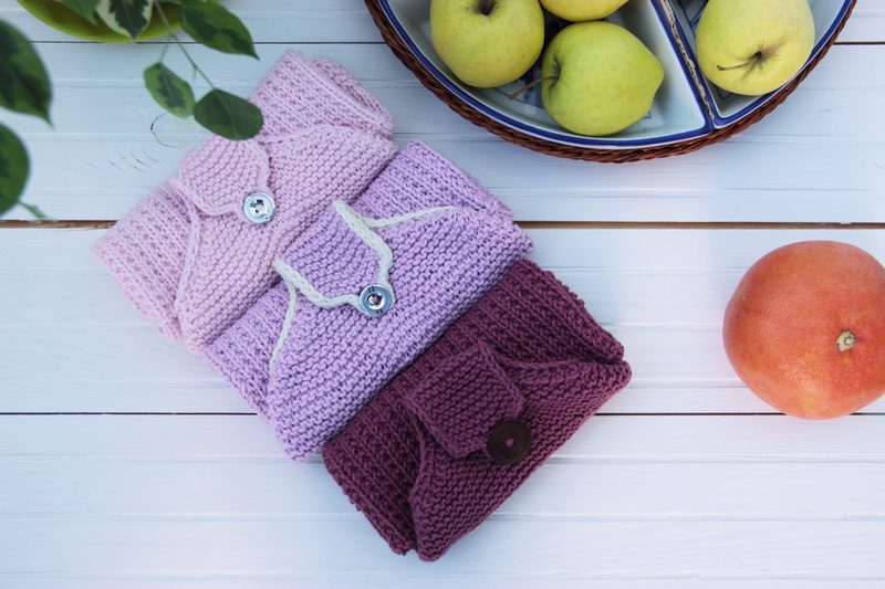The image features three knitted hanging kitchen towels arranged neatly on a white wooden surface. The towels are in shades of light pink, lavender, and deep burgundy, each with a decorative button closure. To the left, there is a small green plant. In the background, a bowl containing several green apples is visible, along with a single orange placed to the right of the towels. The overall composition is bright and colorful, with a cozy, homey feel.