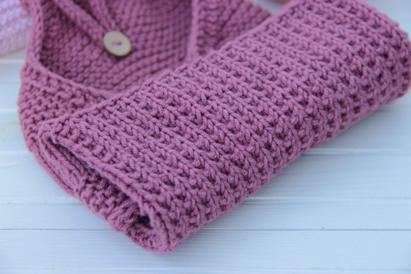 A folded, knitted kitchen towel in a soft pink color is prominently displayed, featuring a textured, woven pattern. The towel has a button closure near the top, and the surface appears smooth and neat. It is set against a light wooden background, which contrasts gently with the pink hue of the towel. The composition emphasizes the towel's shape and texture, with soft natural lighting enhancing the overall appearance.