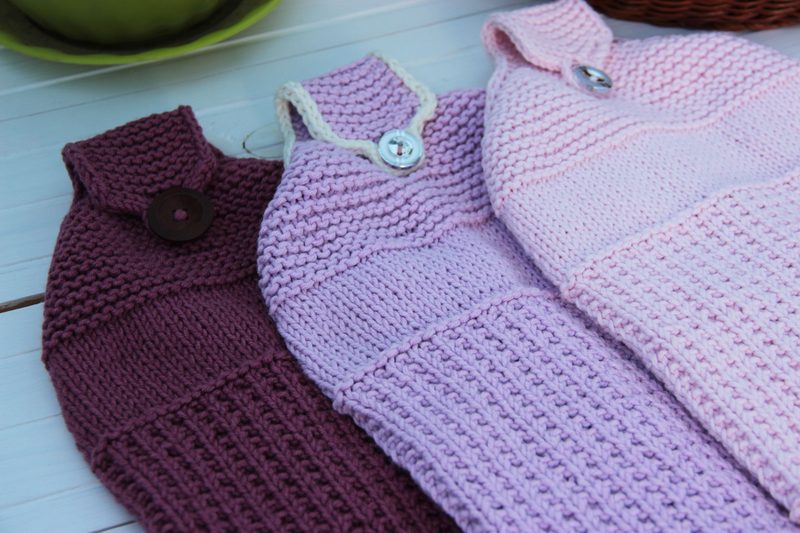 Three hanging kitchen towels made of knitted fabric are displayed in a row. The towels are primarily in shades of pink and purple, featuring different textured patterns. The left towel is in a deep raspberry color with a textured knit and a large dark button. The middle towel has a lighter lavender hue with a striped pattern and a light-colored button. The right towel is in a soft pink shade, also with a striped design. The background includes a hint of a green bowl and a woven basket, adding a