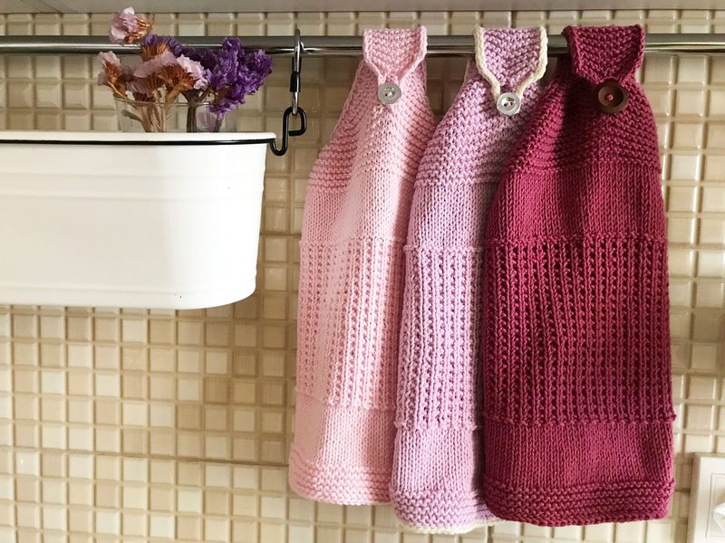 Three knitted hanging kitchen towels are displayed on a horizontal rod. They are arranged in a gradient from light pink on the left to dark burgundy on the right. Each towel features a loop at the top with a button for hanging. Accompanying the towels, a small white metal container holds a bouquet of dried flowers in shades of purple. The background consists of a light-colored tiled wall, adding a subtle texture to the scene.