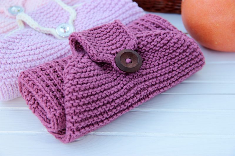 A rolled-up knitted kitchen towel in a rich berry color, secured with a wooden button at the top, is displayed on a light wooden surface. The towel features a textured stitch pattern and is positioned near a round orange and a woven basket in the background. A pink knitted towel is partially visible behind the main towel, adding a soft contrast to the overall arrangement. The lighting is bright and natural, enhancing the colors and textures.