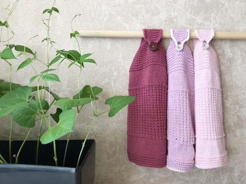 Three knitted kitchen towels hang from a wooden rod, displaying a gradient of colors from dark maroon to light pink. Each towel features a textured pattern with a folded top section that includes buttons for fastening. A green vine plant in a black pot is positioned to the left, complementing the soft tones of the towels against a textured beige wall in the background.