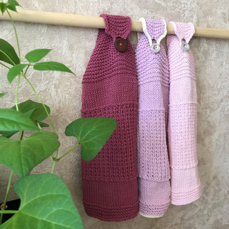 Three hanging knitted kitchen towels are displayed on a wooden rod against a textured light background. The towels are in shades of burgundy, light pink, and soft pink. Each towel features a button closure at the top, with the burgundy towel having a single button and the pink ones having white buttons. A green leafy plant is partially visible in the foreground, adding a natural element to the composition.