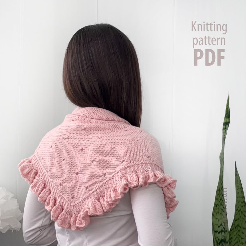 A person with long dark hair is viewed from behind, wearing a delicate pink lace triangle scarf with a frilled edge. The scarf features a textured design and is draped around their shoulders. The background is light-colored, emphasizing the softness of the scarf. On the right side of the image, there is text that reads "Knitting pattern PDF."