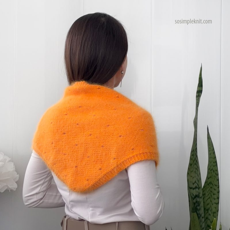 A woman with long brown hair is seen from the back, wearing a vibrant orange lace triangle scarf draped around her shoulders. The scarf features a delicate pattern that is highlighted by the interplay of light and shadow. She is dressed in a white top, and the background includes a white wall and a green plant with elongated leaves, adding a natural element to the scene. The overall setting is bright and minimalistic.