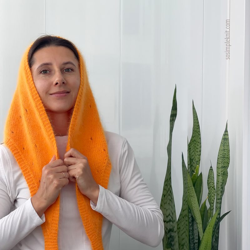 A woman is wearing a bright orange lace triangle scarf draped over her head and shoulders, with her hands partially holding the scarf. She is dressed in a white long-sleeve top and is positioned against a simple white background. To her right, there is a tall green plant with long, pointed leaves, adding a natural element to the scene. The overall composition focuses on the scarf's texture and vibrant color.