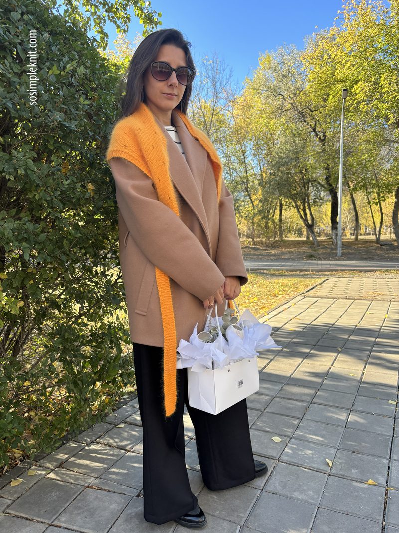 A woman stands outdoors next to a bush, wearing a beige oversized coat and black wide-leg trousers. She has a long orange lace triangle scarf draped around her shoulders and holds a white gift bag filled with tissue paper in one hand. The background features trees with yellow leaves against a clear blue sky, creating a bright autumn setting. The scene is well-lit, emphasizing the colors of her outfit and the scarf.