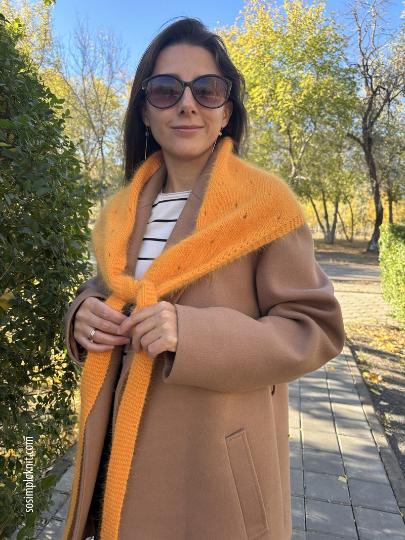 A woman stands outdoors on a stone pathway surrounded by greenery and autumn trees. She wears a beige coat over a black and white striped shirt. Draped around her shoulders is a vibrant orange lace triangle scarf that ties in the front. She is wearing large sunglasses and has shoulder-length dark hair, giving a warm, casual appearance against the bright fall scenery.