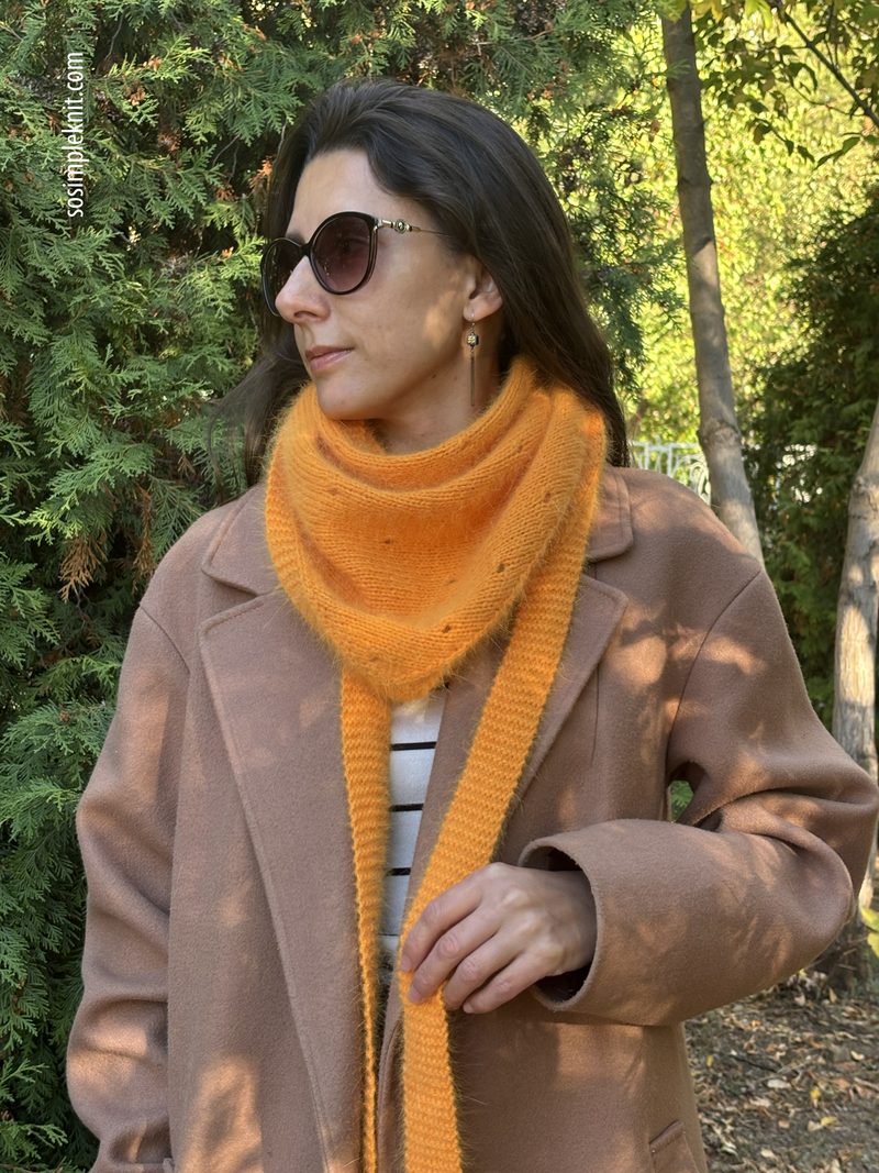 A woman stands outdoors wearing a large orange lace triangle scarf draped around her neck and shoulders, paired with a light brown coat. Her hair is long and dark, partially covering her shoulder, and she wears dark sunglasses. The background features lush greenery, suggesting a natural setting, with soft sunlight illuminating the scene.