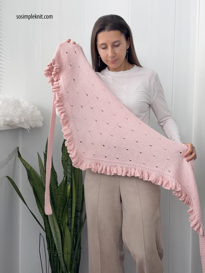 A woman holds a large, pink lace triangle scarf made from knitted fabric, featuring a ruffled edge and small eyelet patterns throughout. She wears a long-sleeved white top and stands in front of a white background, partially obscured by a large potted plant. The overall composition showcases the scarf's texture and design while highlighting the model's gentle pose.