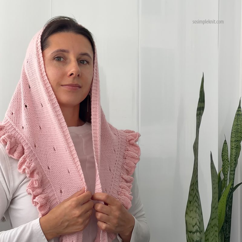 A woman in a light gray long-sleeve shirt is wearing a soft pink lace triangle scarf, featuring ruffled edges. The scarf drapes around her shoulders and frames her face, accentuating her subtle expression. She poses against a bright, white wall, with a tall green plant visible on the right side of the image, adding a touch of nature to the scene. The overall ambiance is calm and elegant, highlighting the scarf's delicate texture and color.