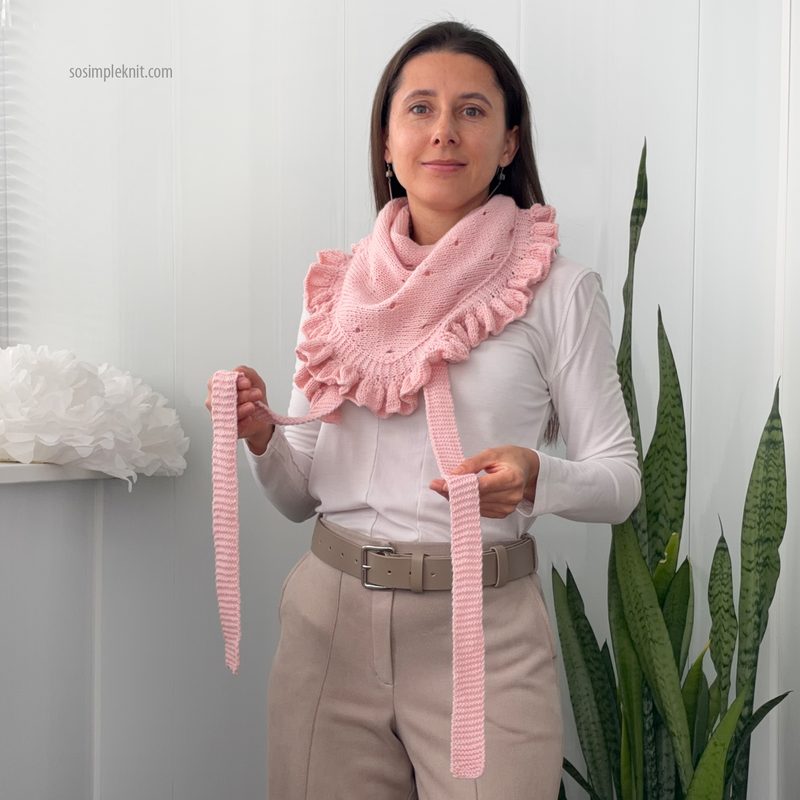 A woman stands in front of a white wall holding a light pink lace triangle scarf, which features a ruffled edge. She wears a long-sleeve white shirt and high-waisted beige pants. The scarf is draped around her shoulders, and she holds the ends, showcasing its length. There are green plants in the background, providing a contrasting natural element.