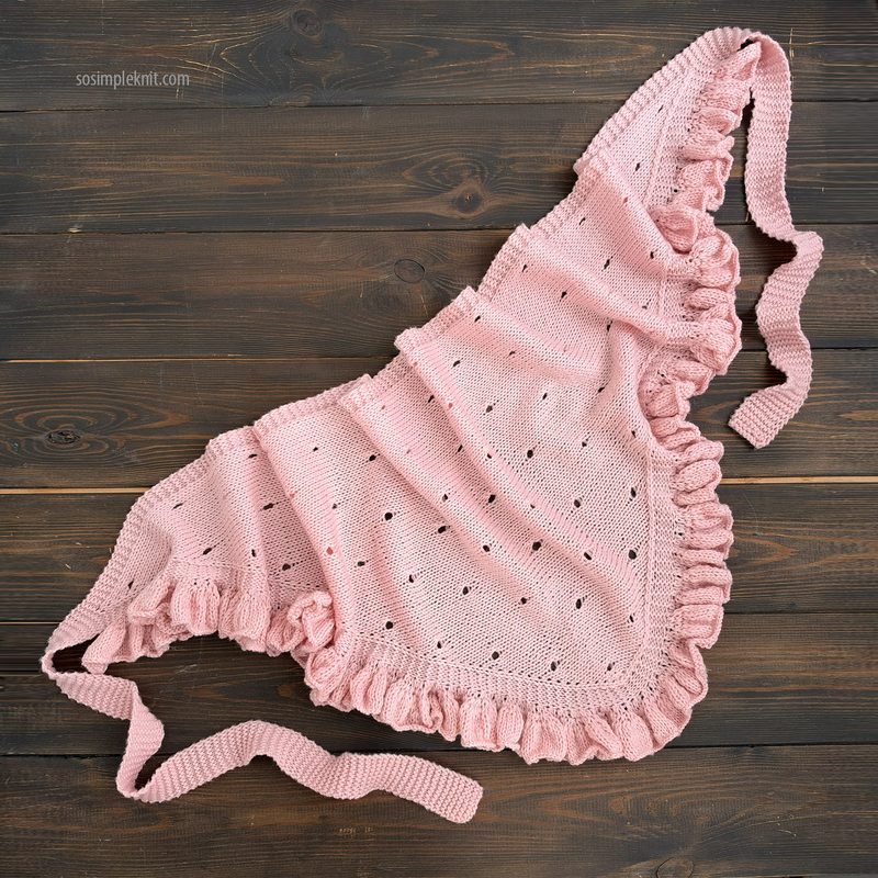 A lace triangle scarf lays flat on a dark wooden surface. The scarf is a soft pink color with a frilled edge and features a delicate, openwork lace pattern throughout. Two elongated ties extend from the tips of the triangle, providing functionality for securing the scarf. The overall design is lightweight and airy, emphasizing a handcrafted appearance.