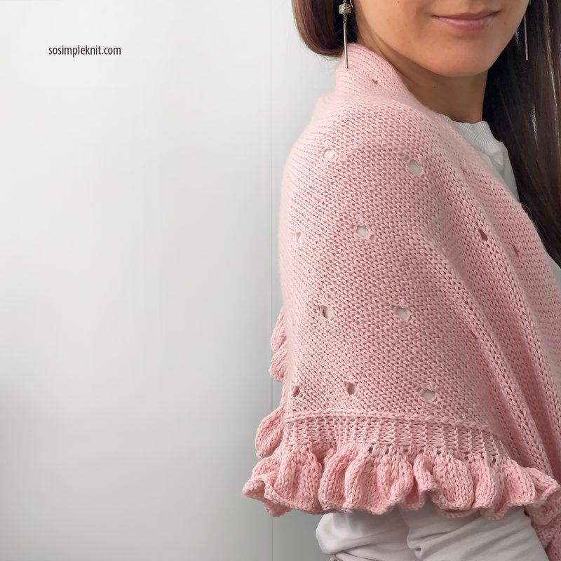 A woman is wearing a light pink lace triangle scarf draped over her shoulders, featuring a textured knit pattern with small eyelet details. The scarf has a scalloped edge at the bottom. The background is a soft white, and the woman has long brown hair, which cascades over one shoulder. She is positioned slightly to the right of the frame, smiling gently, with her face partly turned towards the camera.