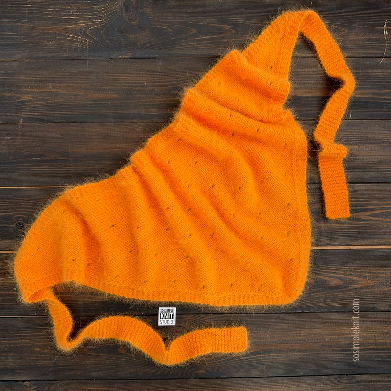 A vibrant orange lace triangle scarf is laid out on a dark wooden surface. The scarf features a delicate, airy texture, showcasing small holes throughout its fabric. It is styled with a cowl neck and has two long ties extending from the edges, creating a relaxed drape. The overall look is soft and inviting, emphasizing the intricate knitting pattern.