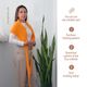 A woman stands in front of a tall green plant, wearing an orange lace triangle scarf draped around her shoulders. She has long brown hair and is dressed in a white top and beige high-waisted trousers. The background is light and neutral, emphasizing the vibrant color of the scarf. To the right, step-by-step instructions are listed in a vertical format, with a decorative knitting-themed element at the top.