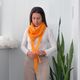 A woman is wearing a vibrant orange lace triangle scarf, elegantly draped around her neck and tied in front. She is dressed in a long-sleeve white shirt and light-colored trousers. The background features a minimalist white wall and a tall, green plant with long leaves, creating a fresh and modern aesthetic. The lighting is bright and soft, highlighting the texture of the scarf.