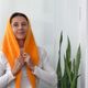 A woman is wearing a bright orange lace triangle scarf draped over her head and shoulders, with her hands partially holding the scarf. She is dressed in a white long-sleeve top and is positioned against a simple white background. To her right, there is a tall green plant with long, pointed leaves, adding a natural element to the scene. The overall composition focuses on the scarf's texture and vibrant color.