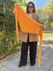 A woman wearing sunglasses holds a vibrant orange lace triangle scarf in an outdoor setting, surrounded by greenery and trees. The scarf is lightweight and features a delicate lace pattern with small holes. The woman is dressed in a beige coat, black pants, and a striped top, standing on a paved path. The lighting suggests a sunny day, enhancing the bright colors of the scarf and the natural surroundings.