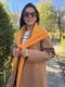 A woman stands outdoors on a stone pathway surrounded by greenery and autumn trees. She wears a beige coat over a black and white striped shirt. Draped around her shoulders is a vibrant orange lace triangle scarf that ties in the front. She is wearing large sunglasses and has shoulder-length dark hair, giving a warm, casual appearance against the bright fall scenery.