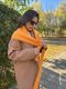 A woman stands outdoors, wearing a light brown coat and large sunglasses. She is draping a vibrant orange lace triangle scarf around her shoulders, with one end held in her hands. The background features green and yellow trees with autumn leaves, and a paved walkway. The scene is well-lit with natural sunlight, creating a warm atmosphere.