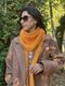A woman stands outdoors wearing a large orange lace triangle scarf draped around her neck and shoulders, paired with a light brown coat. Her hair is long and dark, partially covering her shoulder, and she wears dark sunglasses. The background features lush greenery, suggesting a natural setting, with soft sunlight illuminating the scene.
