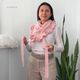 A woman stands in front of a white wall holding a light pink lace triangle scarf, which features a ruffled edge. She wears a long-sleeve white shirt and high-waisted beige pants. The scarf is draped around her shoulders, and she holds the ends, showcasing its length. There are green plants in the background, providing a contrasting natural element.