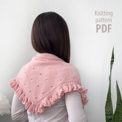 A person with long dark hair is viewed from behind, wearing a delicate pink lace triangle scarf with a frilled edge. The scarf features a textured design and is draped around their shoulders. The background is light-colored, emphasizing the softness of the scarf. On the right side of the image, there is text that reads "Knitting pattern PDF."